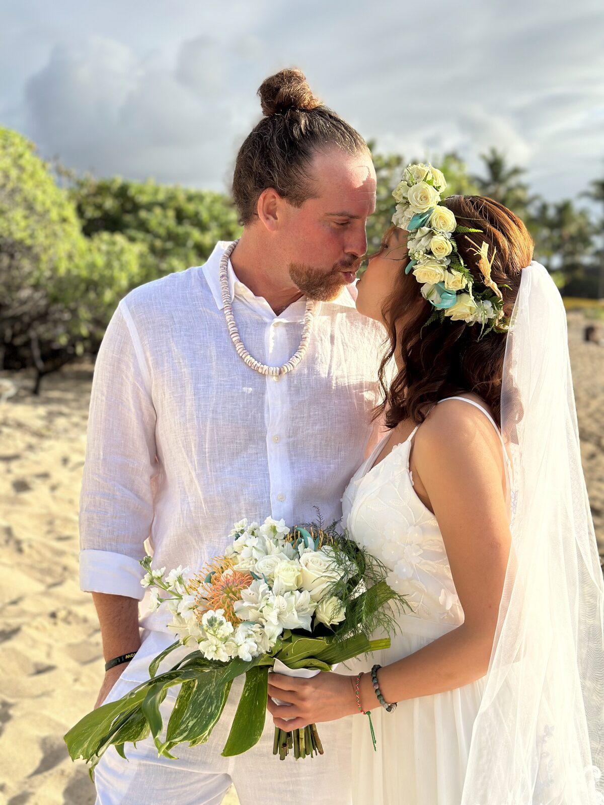 Bride and groom kissing on a Hawaiian beach, groom wearing a puka shell necklace with white linen